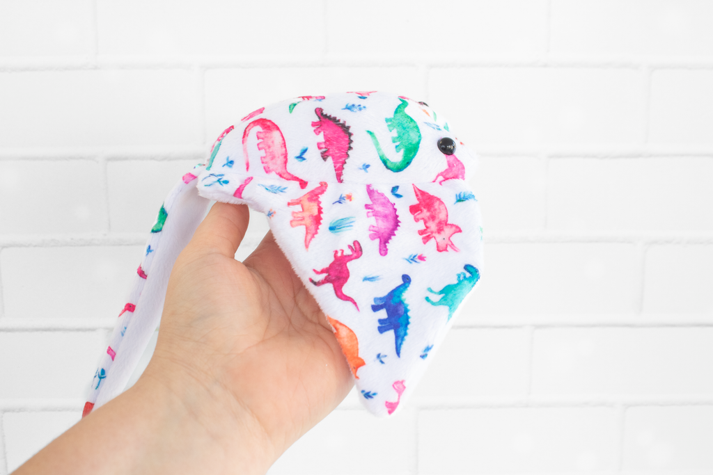 A handmade plush stingray ray featuring colorful dinosaur-themed fabric. The fabric displays vibrant dinosaurs in shades of pink, green, blue, and orange on a white background. The plush has black, round safety eyes and is displayed on a light wooden surface.