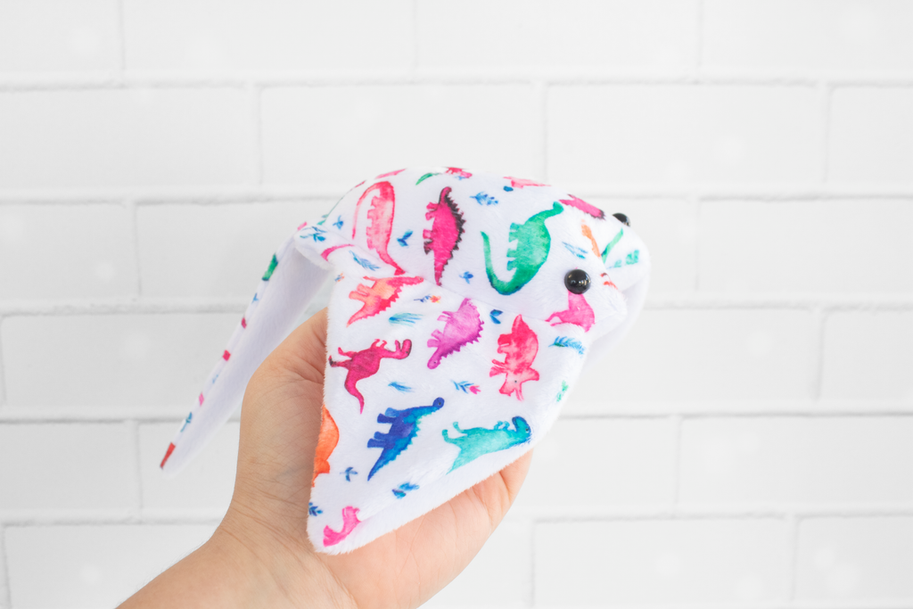 A handmade plush stingray ray featuring colorful dinosaur-themed fabric. The fabric displays vibrant dinosaurs in shades of pink, green, blue, and orange on a white background. The plush has black, round safety eyes and is displayed on a light wooden surface.