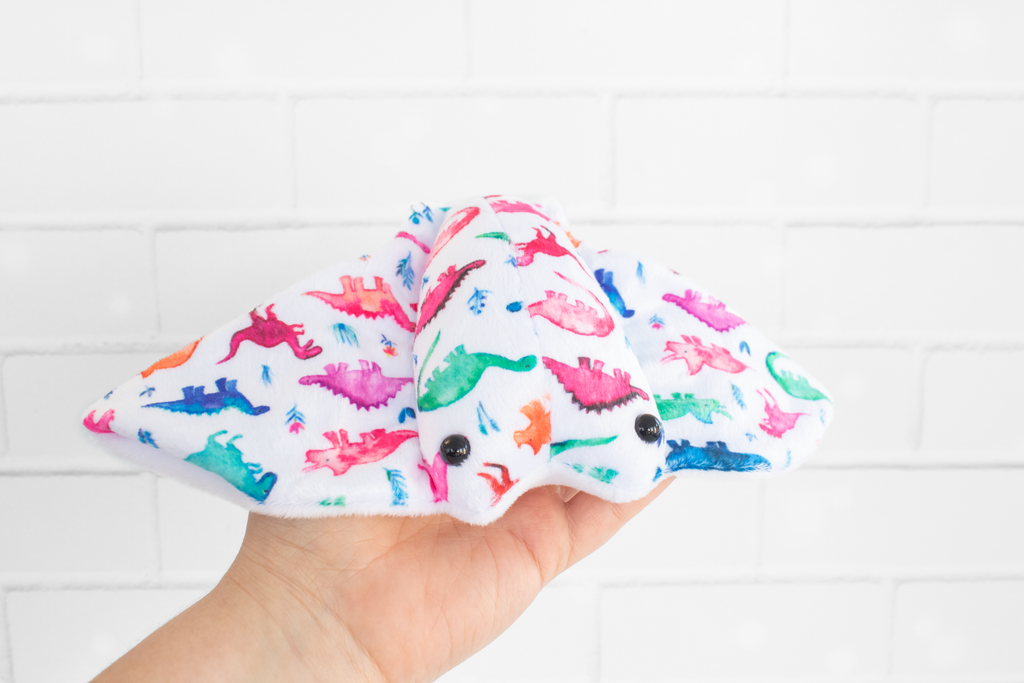 A handmade plush stingray ray featuring colorful dinosaur-themed fabric. The fabric displays vibrant dinosaurs in shades of pink, green, blue, and orange on a white background. The plush has black, round safety eyes and is displayed on a light wooden surface.