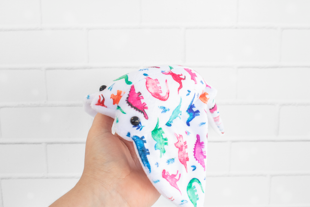 A handmade plush stingray ray featuring colorful dinosaur-themed fabric. The fabric displays vibrant dinosaurs in shades of pink, green, blue, and orange on a white background. The plush has black, round safety eyes and is displayed on a light wooden surface.