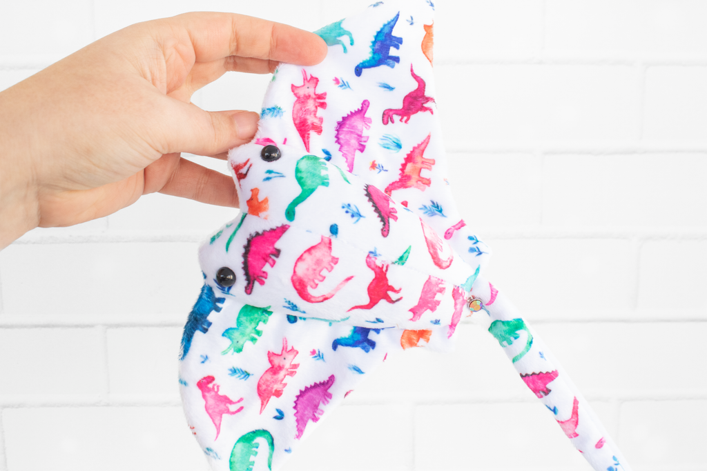 A handmade plush stingray ray featuring colorful dinosaur-themed fabric. The fabric displays vibrant dinosaurs in shades of pink, green, blue, and orange on a white background. The plush has black, round safety eyes and is displayed on a light wooden surface.