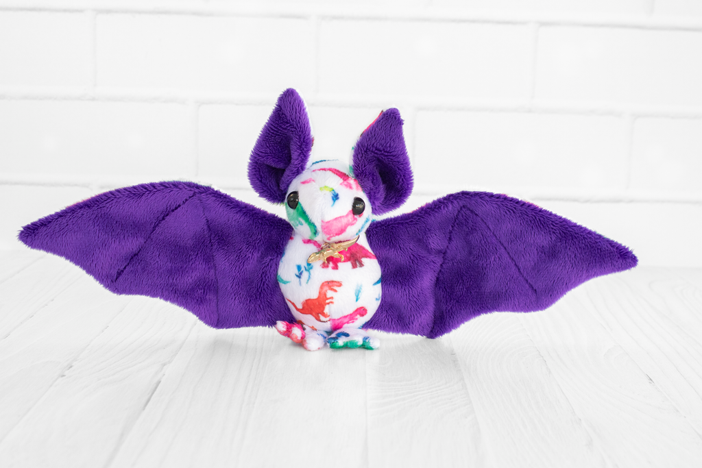 A handmade plush bat with colorful dinosaur-themed fabric on its body. The bat has purple wings and ears and black safety eyes. There is a gold colored dinosaur charm at its neck. It sits on a white wooden surface with a white brick background.