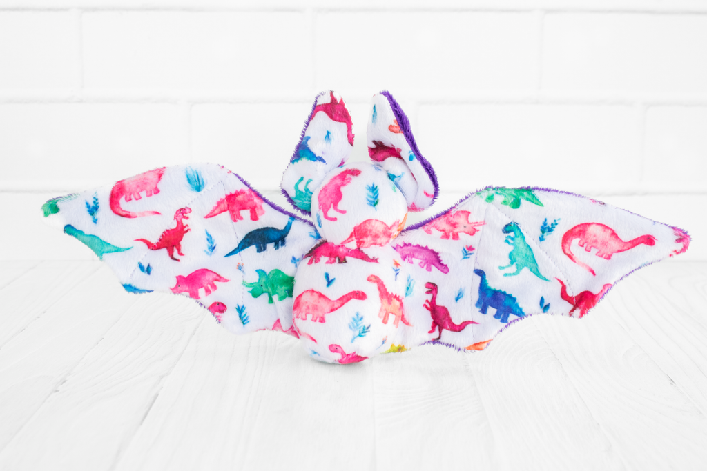 The back view of a handmade bat plush toy. It shows the fabric print which is multicolored dinosaurs on a white background.