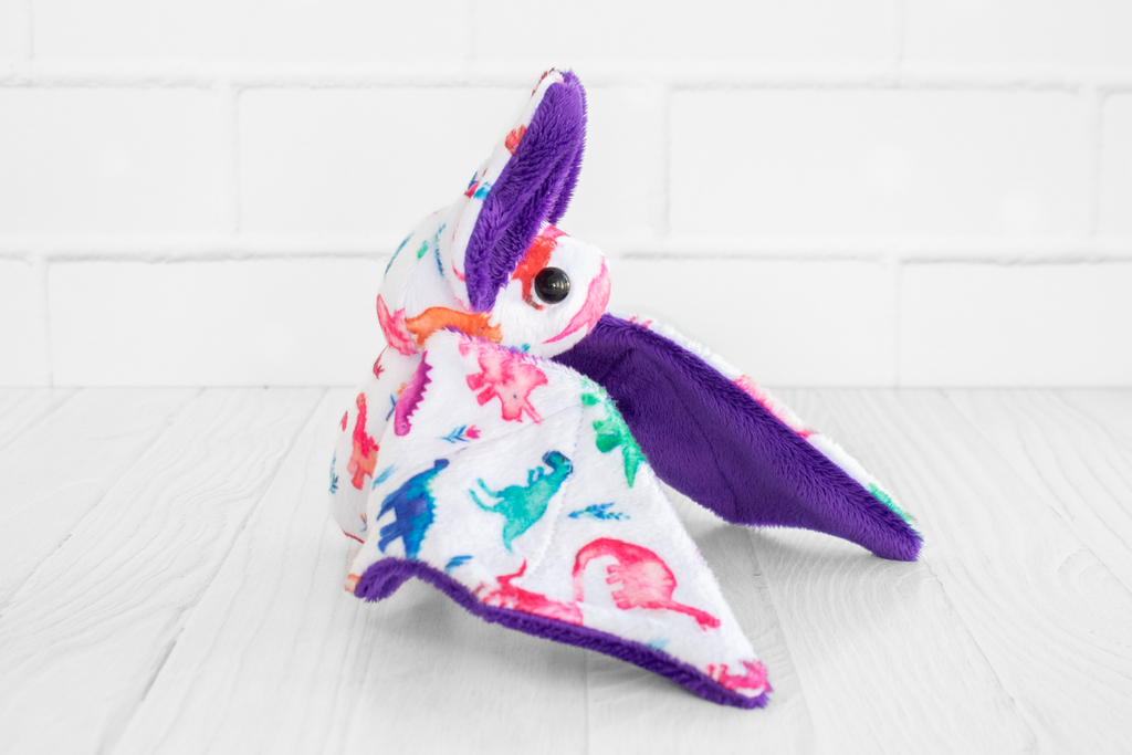 A handmade plush bat with colorful dinosaur-themed fabric on its body. The bat has purple wings and ears and black safety eyes. There is a plastic translucent pink gummy bear charm at its neck. It sits on a white wooden surface with a white brick background.