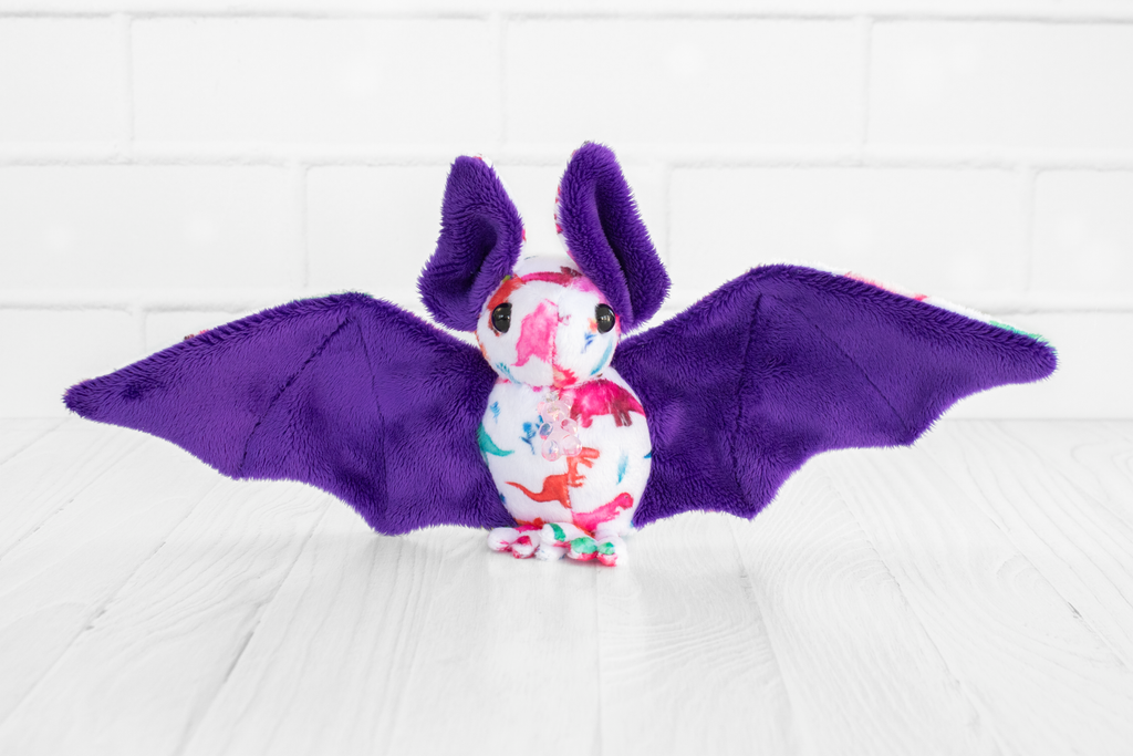 A handmade plush bat with colorful dinosaur-themed fabric on its body. The bat has purple wings and ears and black safety eyes. There is a plastic translucent pink gummy bear charm at its neck. It sits on a white wooden surface with a white brick background.