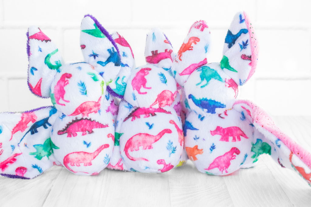 The back of 3 handmade bat plush toys showing off the fabric print which features multicolored dinosaurs on a white background. They sit on a white wood surface with a white brick background.