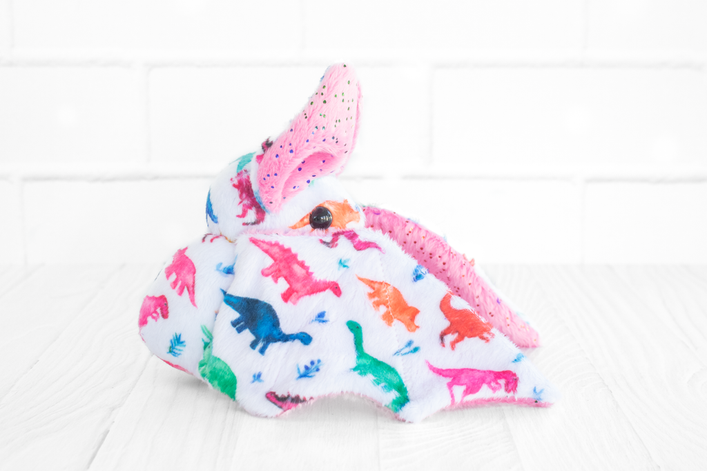 A handmade plush bat with colorful dinosaur-themed fabric on its body. The bat has pink wings and ears with multicolored sparkles and round, black safety eyes. There is a translucent pink plastic gummy bear charm at its neck. It sits on a white wooden surface with a white brick background.