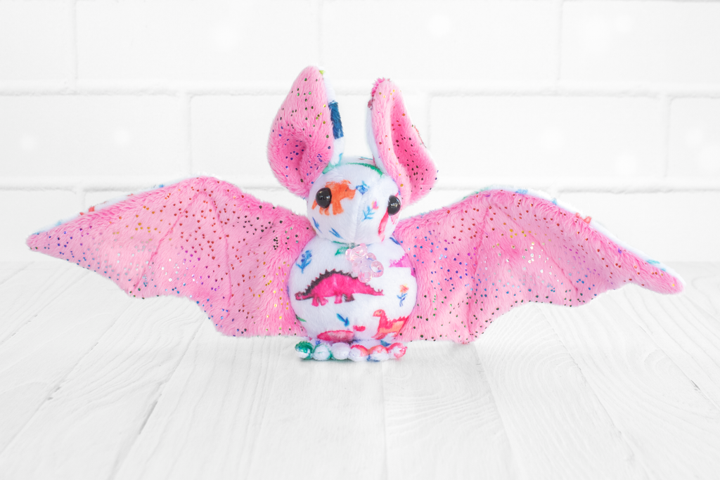 A handmade plush bat with colorful dinosaur-themed fabric on its body. The bat has pink wings and ears with multicolored sparkles and round, black safety eyes. There is a translucent pink plastic gummy bear charm at its neck. It sits on a white wooden surface with a white brick background.