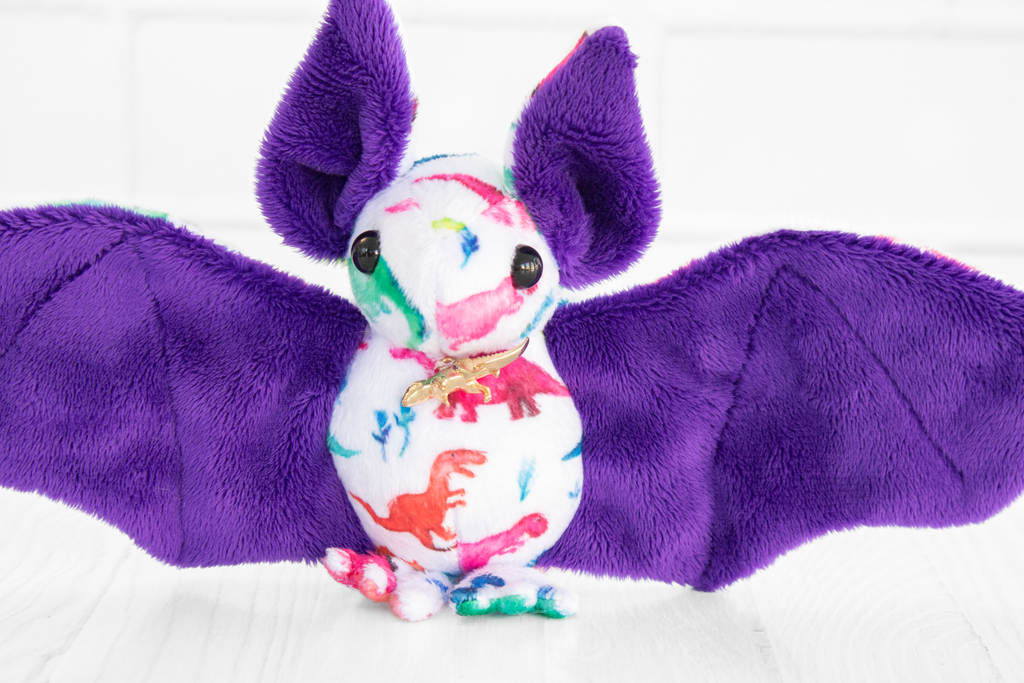 A handmade plush bat with colorful dinosaur-themed fabric on its body. The bat has purple wings and ears and black safety eyes. There is a gold dino charm at its neck. It sits on a white wooden surface with a white brick background.