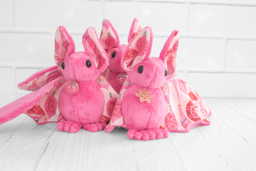 A group of pink bat plushies. The wing and ear fabric shows red and pink christmas ornaments. Some have glass globes with little gems at their neck and some have gold snowflakes with pink glitter inside. They are sitting on a white wood surface with white brick background. 