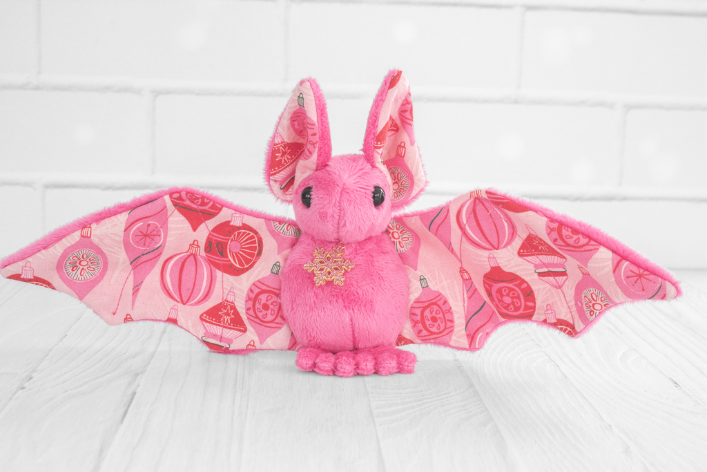 A pink plush bat with wings spread out. The wing fabric features a festive pattern of red and pink Christmas ornaments. The plush bat is standing on a white wooden surface with a white brick background. It is adorned with a small golden snowflake charm on its chest.