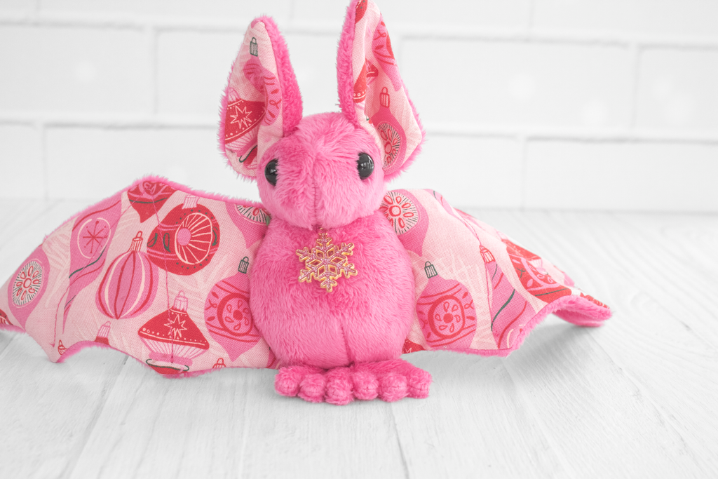 A pink plush bat with wings spread out. The wing fabric features a festive pattern of red and pink Christmas ornaments. The plush bat is standing on a white wooden surface with a white brick background.