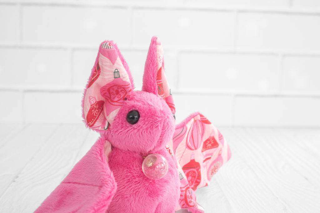 A pink plush bat zoomed in to show mostly the face which has solid black, round safety eyes. The wing fabric features a festive pattern of red and pink Christmas ornaments. The plush bat is sitting on a white wooden surface with a white brick background.