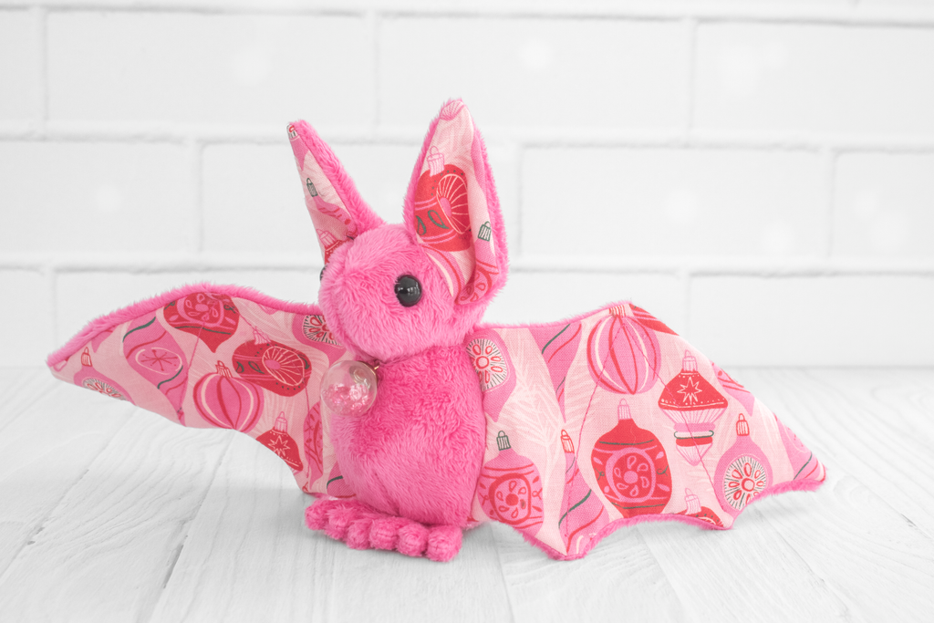 The pink plush bat facing forward with its wings slightly closed. It is adorned with a small golden snowflake charm on its chest. The wings and ears feature the red and pink ornament pattern, adding a holiday-themed touch.