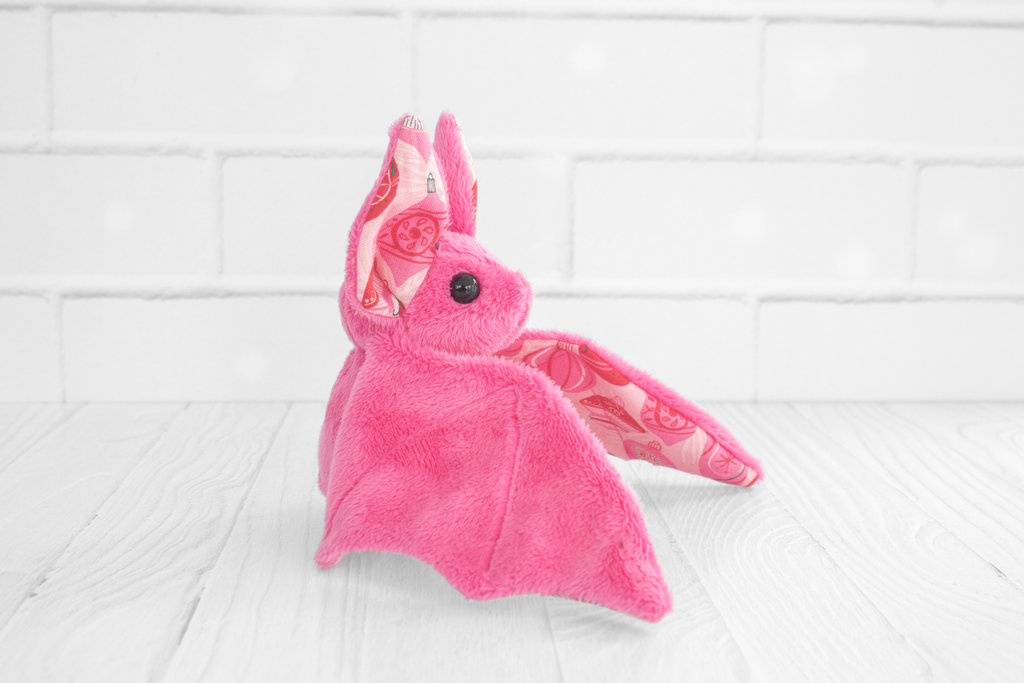 A side profile view of the pink plush bat. Its ears and wings display the same festive ornament pattern. It is positioned on the same white wooden surface with a brick wall backdrop.