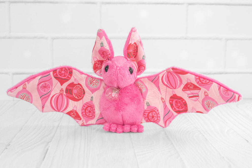 A pink plush bat with wings spread out. The wing fabric features a festive pattern of red and pink Christmas ornaments. The plush bat is standing on a white wooden surface with a white brick background.