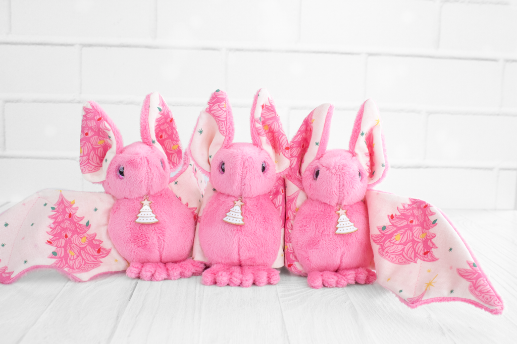 3 handmade pink bat plushies with vintage christmas tree print on their wings and white christmas tree charms on their necks. Their eyes are pink and sparkly with snowflakes