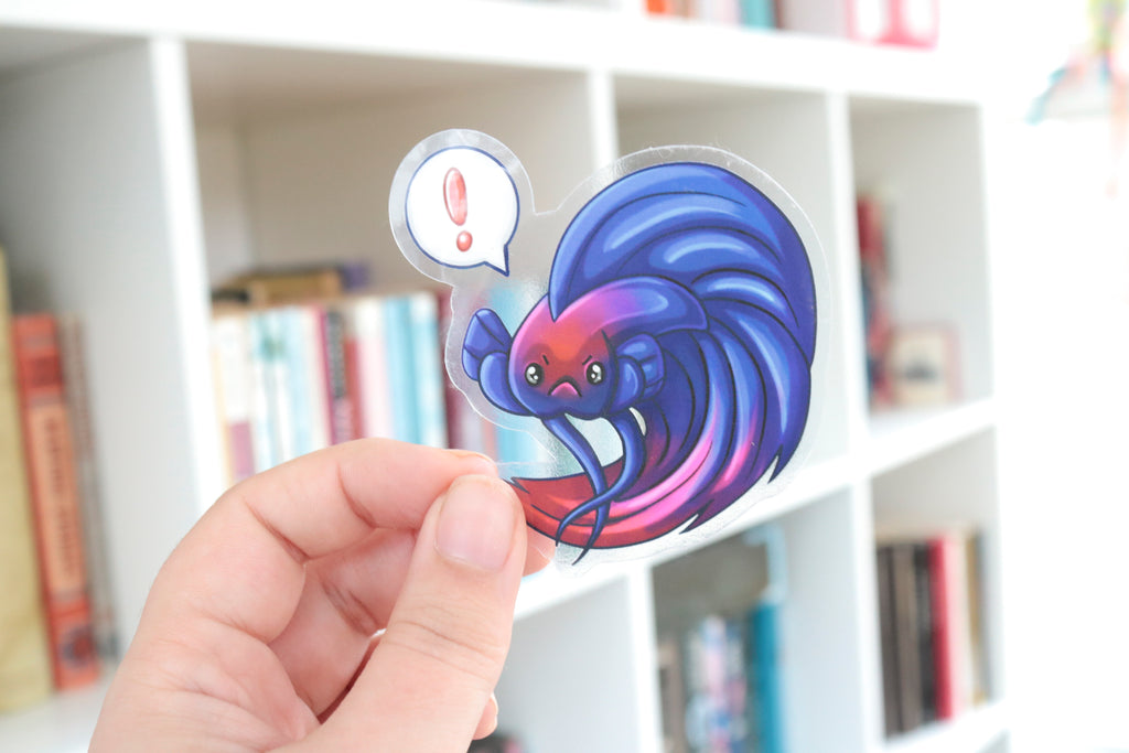 Image shows a hand holding a sticker over a shelf of books. The sticker is transparent around the image printed on it, allowing the bookshelf to show through. The image on the sticker itself is a royal blue betta fish with maroon red color gradients towards the face and end of the tail with a floating speech bubble over its head that has a red exclamation point in the speech bubble.